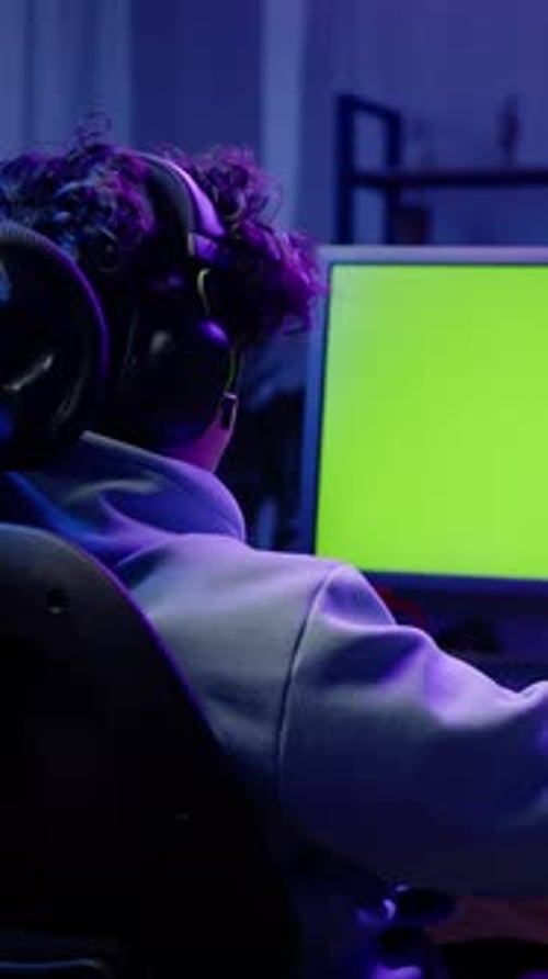 Rear View Young Gamer in Headphones Plays Online Video Game on Laptop with Green Chromakey Screen