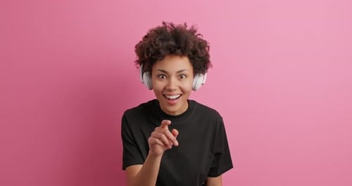 Smiling Woman Points Finger Wearing Headphones Against Pink