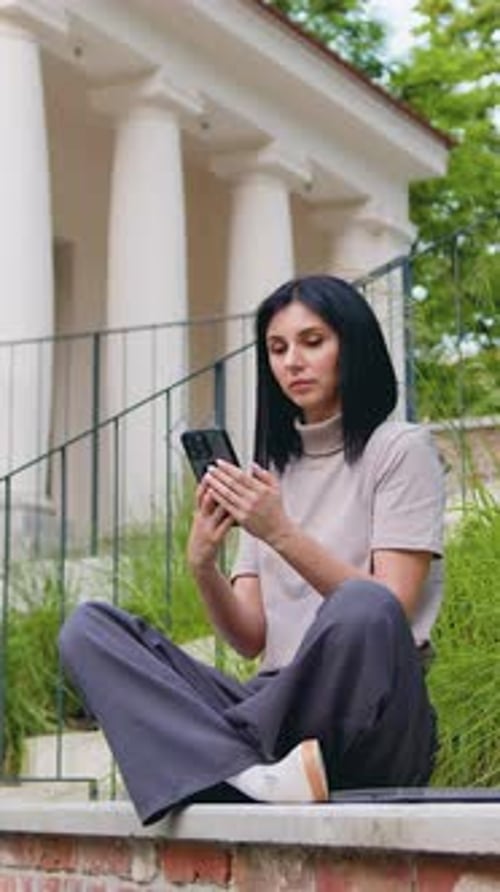 Young Caucasian Woman Use Mobile Phone While Sitting on the Bench Near Modern Old Building in the