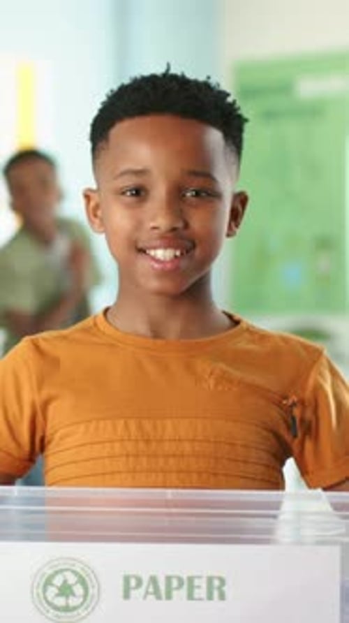 Vertical video: Camera focusing, boy smiling behind PAPER bin in class in tan tee for recycle demo
