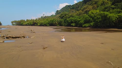 Woman spinning in white dress on wide sandy beach with lush forest on shore in Costa Rica
