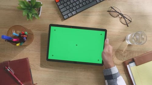 Top down view of man's hand holding a mock up green screen tablet on a desk