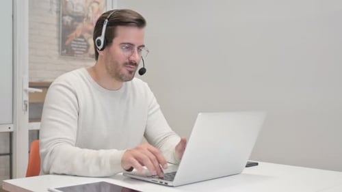 Young Man with Headset Doing Video Chat on Laptop in Call Center