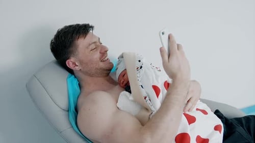 Father Cradling Newborn Baby in Hospital, Looking at Phone