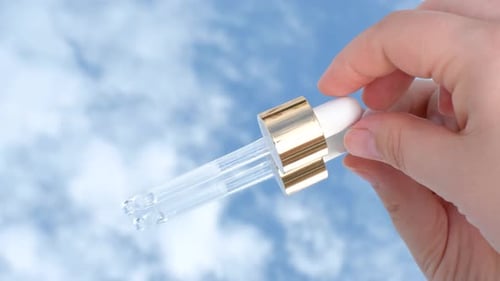 Female hand puts a pipette with serum on blue background with clouds close-up.