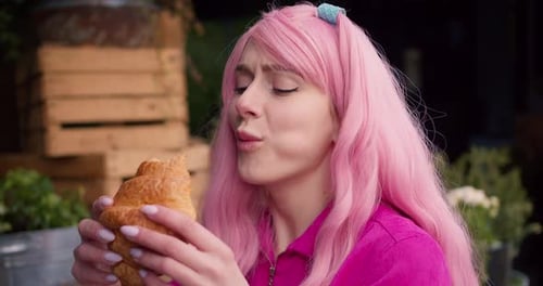 Closeup Shot of a Girl with Pink Hair Tasting a Delicious Croissant in a Street Cafe Happy Summer