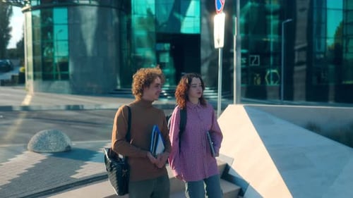 Two College Students Walking with Books Near Modern Glass Campus Building