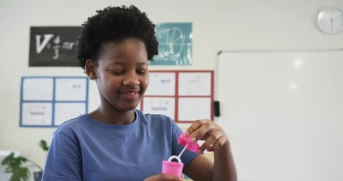 African teen female unscrewing pink bubble bottle dipping wand blowing bubbles in classroom for fun