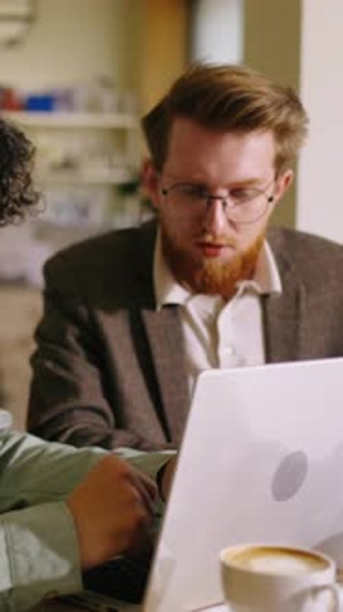 Young Man with Curly Hair Working on a Laptop at a Cozy Cafe Table with Coffee and Pastry Focusing