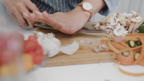 Midsection of biracial woman in apron preparing meal, crushing garlic clove in kitchen, slow motion