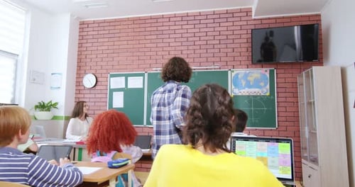 Rückseite eines kaukasischen Teenagers, der im Klassenzimmer vom Schreibtisch steht und zur Tafel geht, Lehrerin