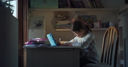 Authentic shot of little girl pupil is writing in notebook at desk while having online lecture wit