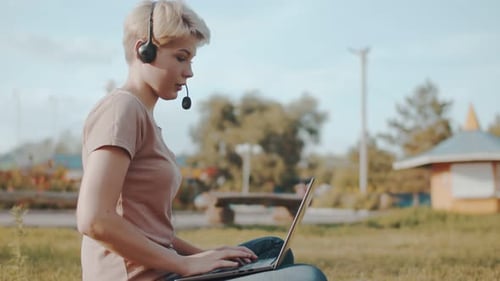 Side View of a Female with Headset is Sitting Park on Grass with Laptop and Says Something Into