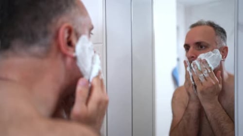 Man applies shaving cream in bathroom mirror