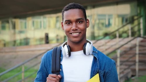 A Smiling Student in Stylish Headphones with a Stack of Books is Seen on a Vibrant College Campus