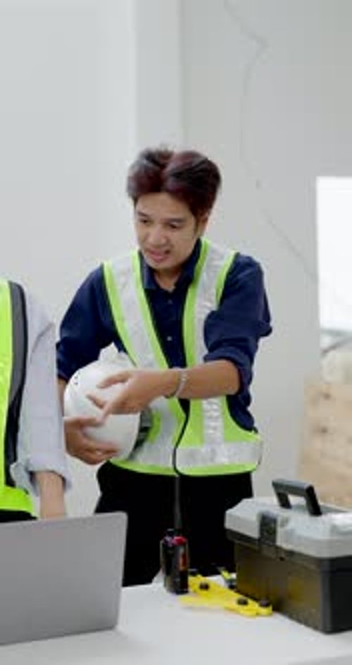 Asian female worker holds white helmet while checking laptop near measuring tool, supporting