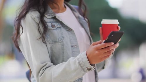 Woman Holding Coffee Looks at Smartphone in City