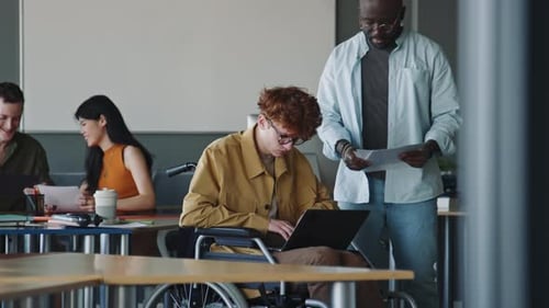 Young Man in Wheelchair Using Laptop and Talking with Coworker in Office