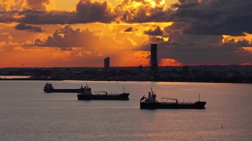 Aerial drone view of multiple oil tankers anchored near an industrial port at sunset with cranes and