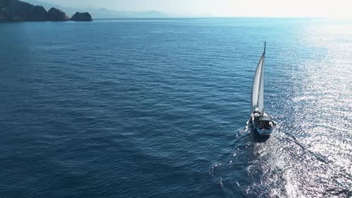 Aerial View Real Time of White Sailboat Floating on Sea Near Rocky Cliff and Town Located on Slope