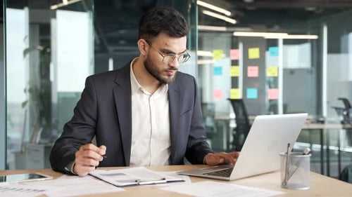 Busy businessman in formal suit making financial calculations using laptop, taking notes at workplac