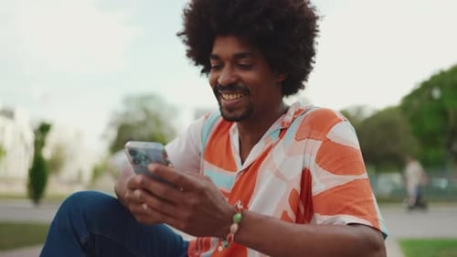 Close-up of young African American man wearing shirt sitting on park bench using his smartphone.