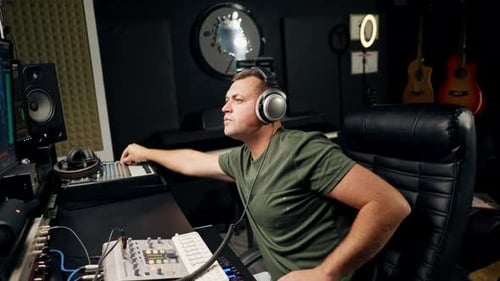 a focused male sound engineer wearing headphones sits at a mixing desk recording new track