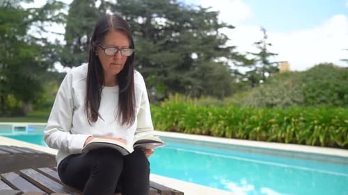 Adult Woman With Eyeglasses Reading Book And Sitting By The Poolside. medium shot