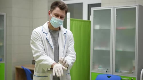 A Male Medical Worker Wearing a White Coat and a Mask Puts on Medical Pants and Prepares for Work