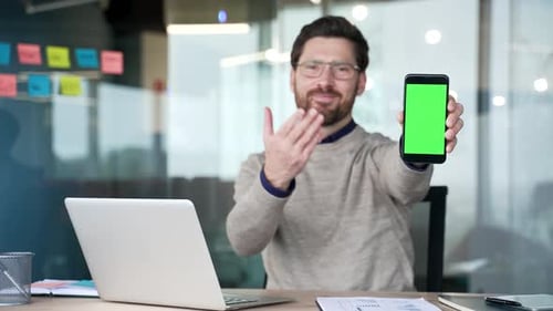 Smiling businessman showing smartphone with green screen while sitting in office.