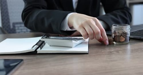 Woman putting coins into glass jar and using calculator at wooden table indoors, closeup
