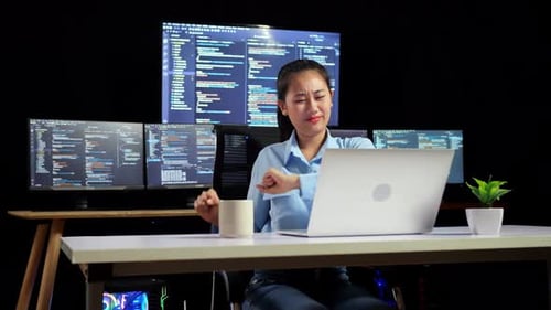 Asian Female Programmer Stretching While Writing Code By A Laptop Using Multiple Monitors