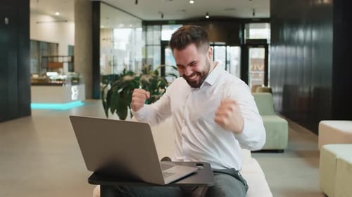 Caucasian Bearded Businessman Working on Laptop Shocked By Sudden Victory in Modern Hotel Lobby