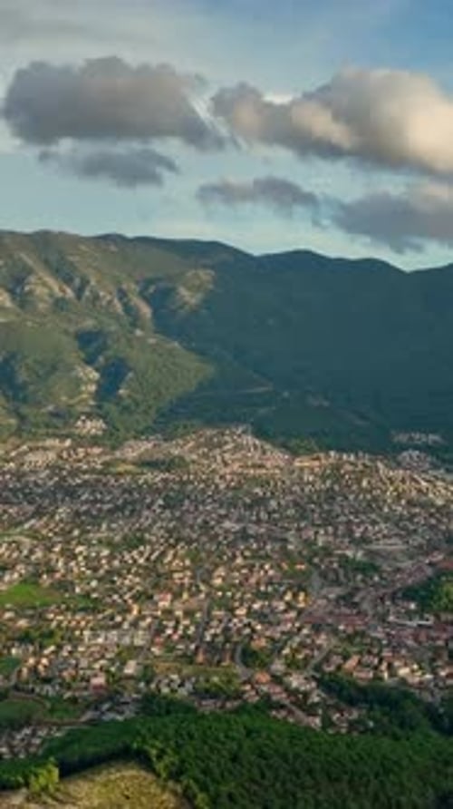 Aerial view of a sprawling town nestled at the base of lush green mountains under a cloudy sky