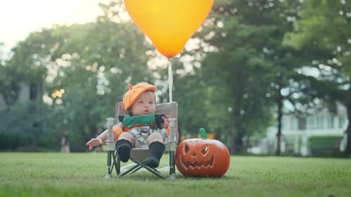Cute small baby girl sitting near pumpkin with orange balloon in park outdoor.