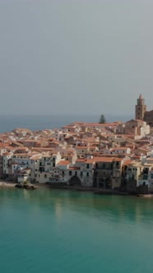 Flying into the Cefalu town above Mediterranean sea. Vertical aerial drone shot of sandy beach town