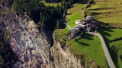small alpine village on the edge of a rocky wall, green grass fiald and pine forest, aerial shot