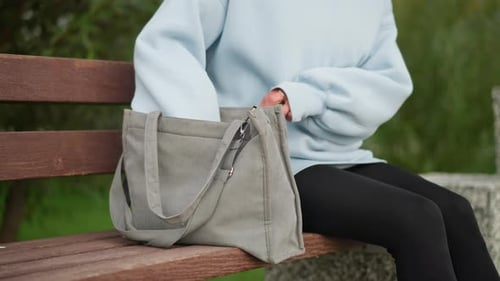 Señora sentada en un banco con ropa casual sacando una botella de agua del bolso en un entorno exterior