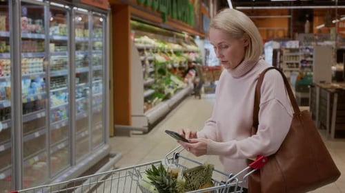 Blonde Mature Woman using Phone in Supermarket Checking Shopping List