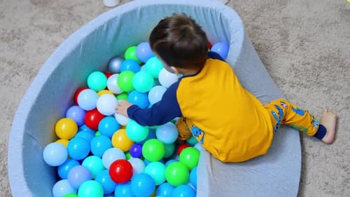 Kid in yellow pajama climbs into the dry pool. Baby boy sits into the colorful balls. Top view.