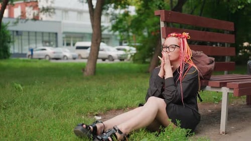 Young Woman Sitting on Grass Near Bench Wearing Glasses and Pink Dreadlocks Hands Pressed to Face