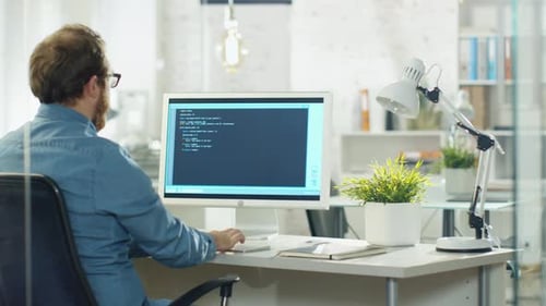 Young Bearded Developer Writes Code on His Desktop Computer. He Works in a Modern Office.