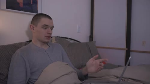 Young Man Sitting On Bed Using A Laptop, Waving, And Talking To His Colleague. - close up shot