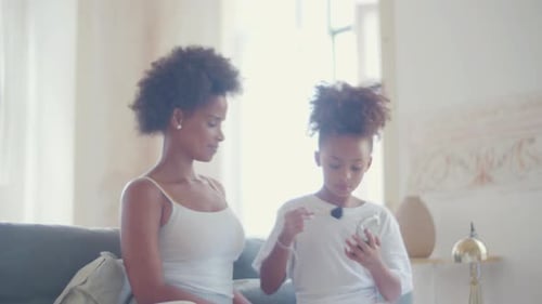 Cute Girl Doing Makeup on Her Mother Sitting on Couch Indoors