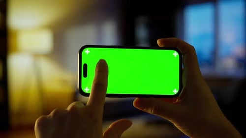Hand Holding Modern Smartphone with Bright Green Chroma Key Screen in Cozy Dimly Lit Living Room