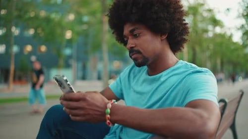 man in blue t-shirt sitting on park bench using his smartphone