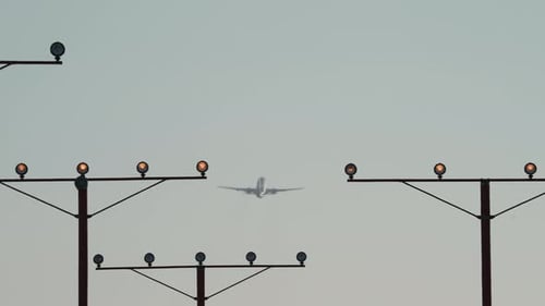 An airliner climbs out after takeoff in the distance, framed by runway approach lights.