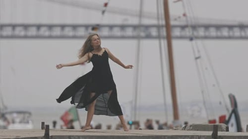 An Adult Woman in Black Dress Dancing on the Pier with a Bridge on the Background and a Yacht