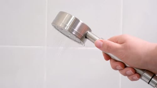 Female hand holding white shower head on white tile background close-up, side view.