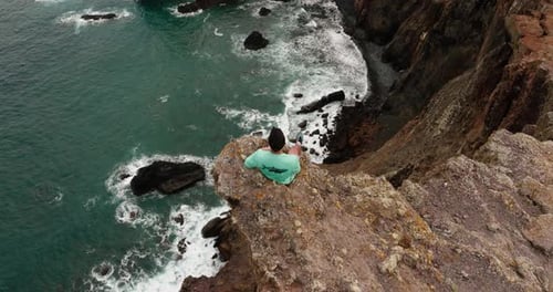 Aerial tilt-up reveals a man sitting on a cliff edge overlooking the dramatic coastline of Ponta de